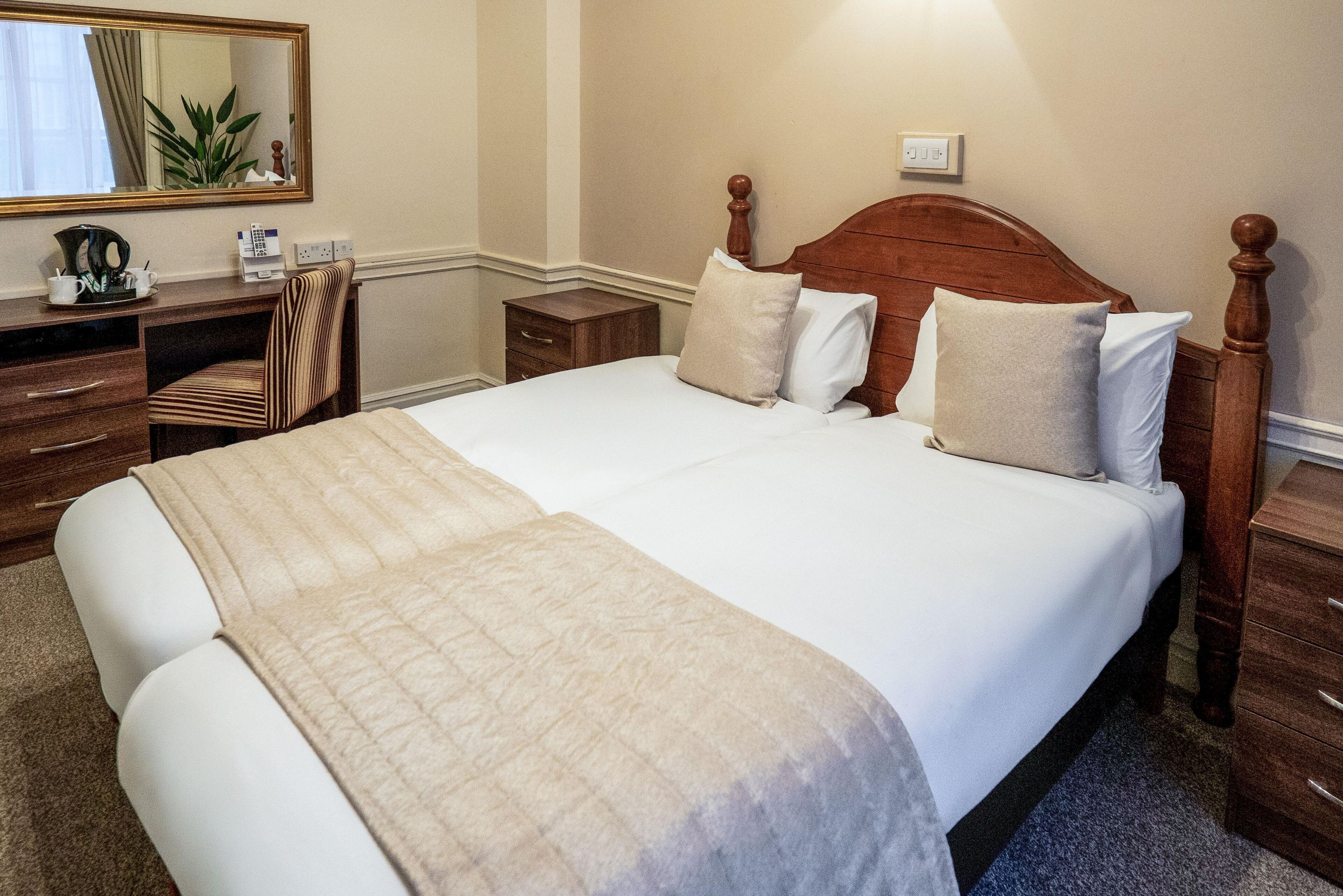 A neatly made bed with a wooden headboard, a bedside table, a chair, and a desk with a mirror and some items on it.