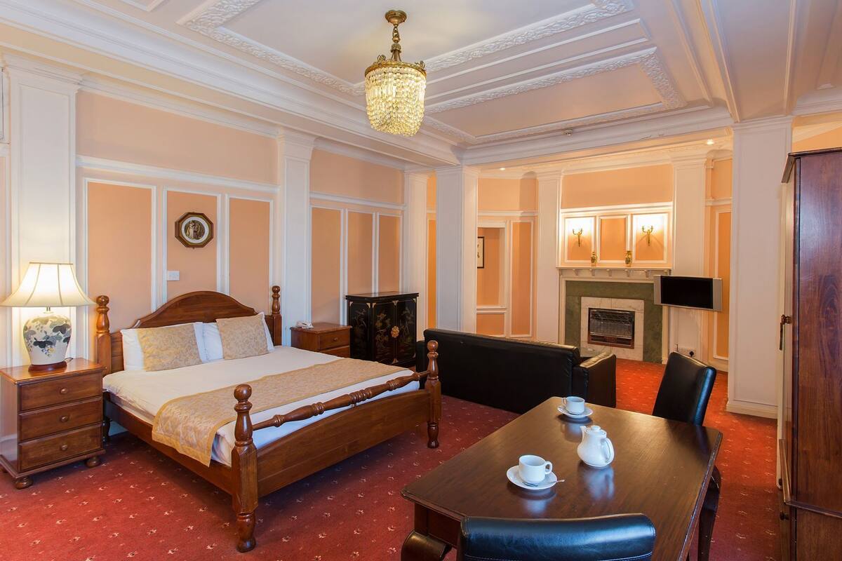 Habitación de hotel con cama, mesita de noche, lámpara y zona de comedor con mesa y sillas.