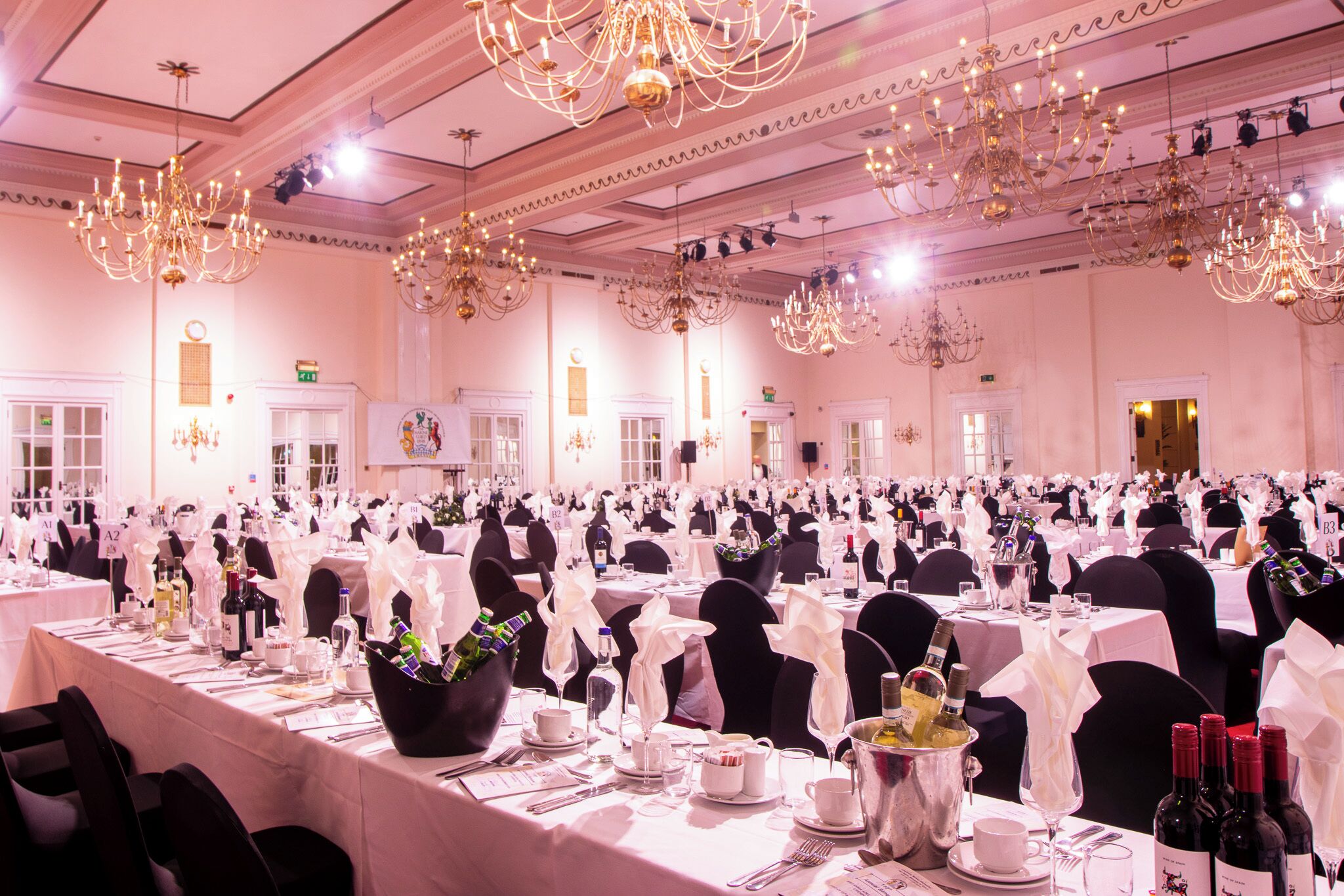 A banquet hall with multiple tables set for a formal event, featuring chandeliers and white tablecloths.
