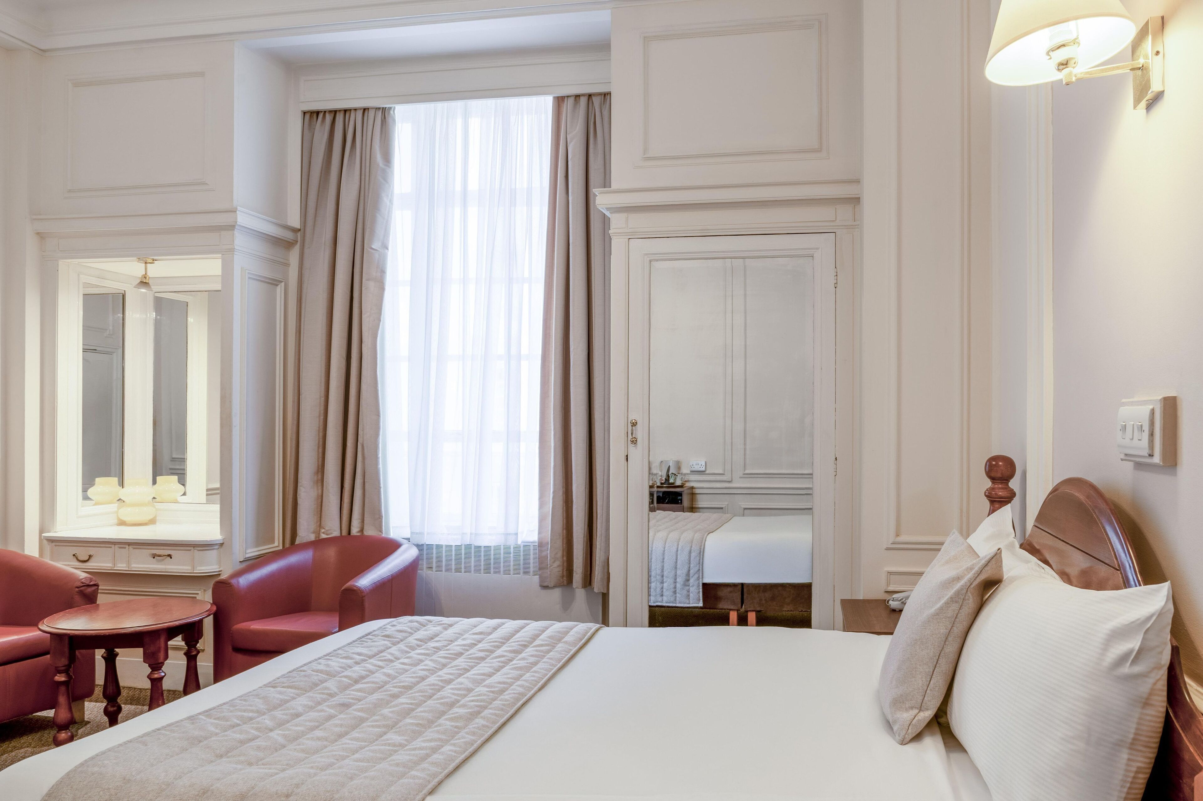 A hotel room with a large bed, a sitting area with a table and chairs, a vanity with a mirror, and a window with curtains.