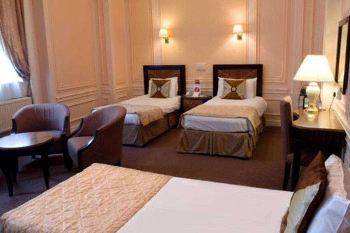 Habitación de hotel con dos camas individuales, un sofá, una mesita pequeña y una mesita de noche con lámpara.