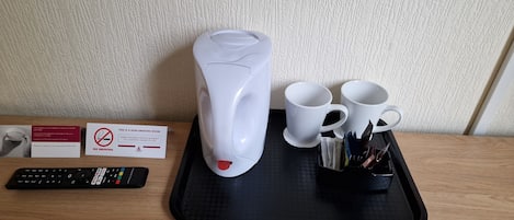 Señal de habitación para no fumadores, una tetera eléctrica blanca, dos tazas blancas, un control remoto y una bandeja con bolígrafos y un cuaderno.