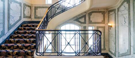 Una imponente escalera con barandilla de metal negro, paredes de mármol y alfombra estampada.