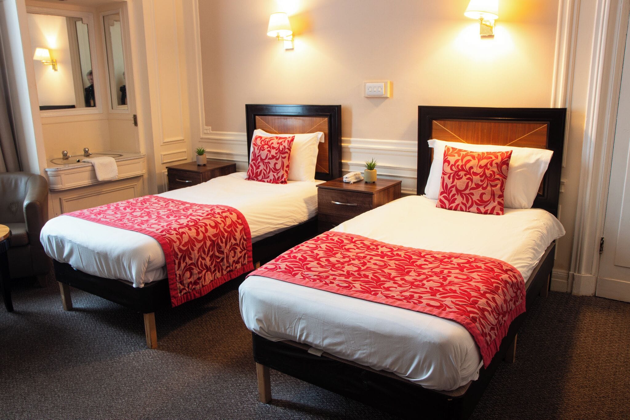 Habitación de hotel con dos camas, cada una con cabecero de madera, ropa de cama blanca y mantas decorativas estampadas en rojo. Se observan mesitas de noche con lámparas, un espejo y una planta pequeña.