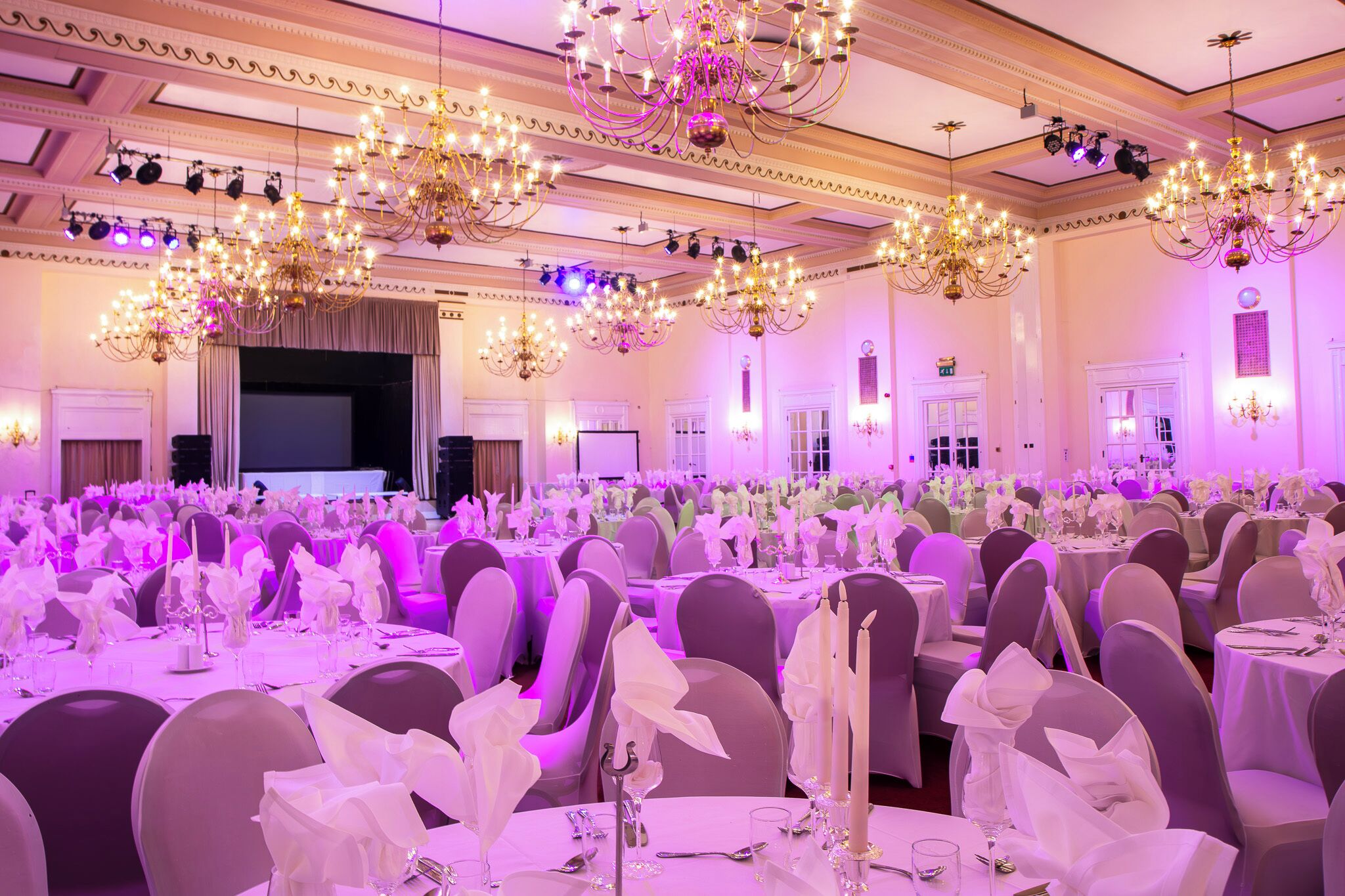 A banquet hall with rows of round tables, each set with white tablecloths, chairs, and floral centerpieces.
