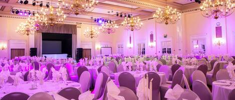 A banquet hall with rows of round tables, each set with white tablecloths, chairs, and floral centerpieces.