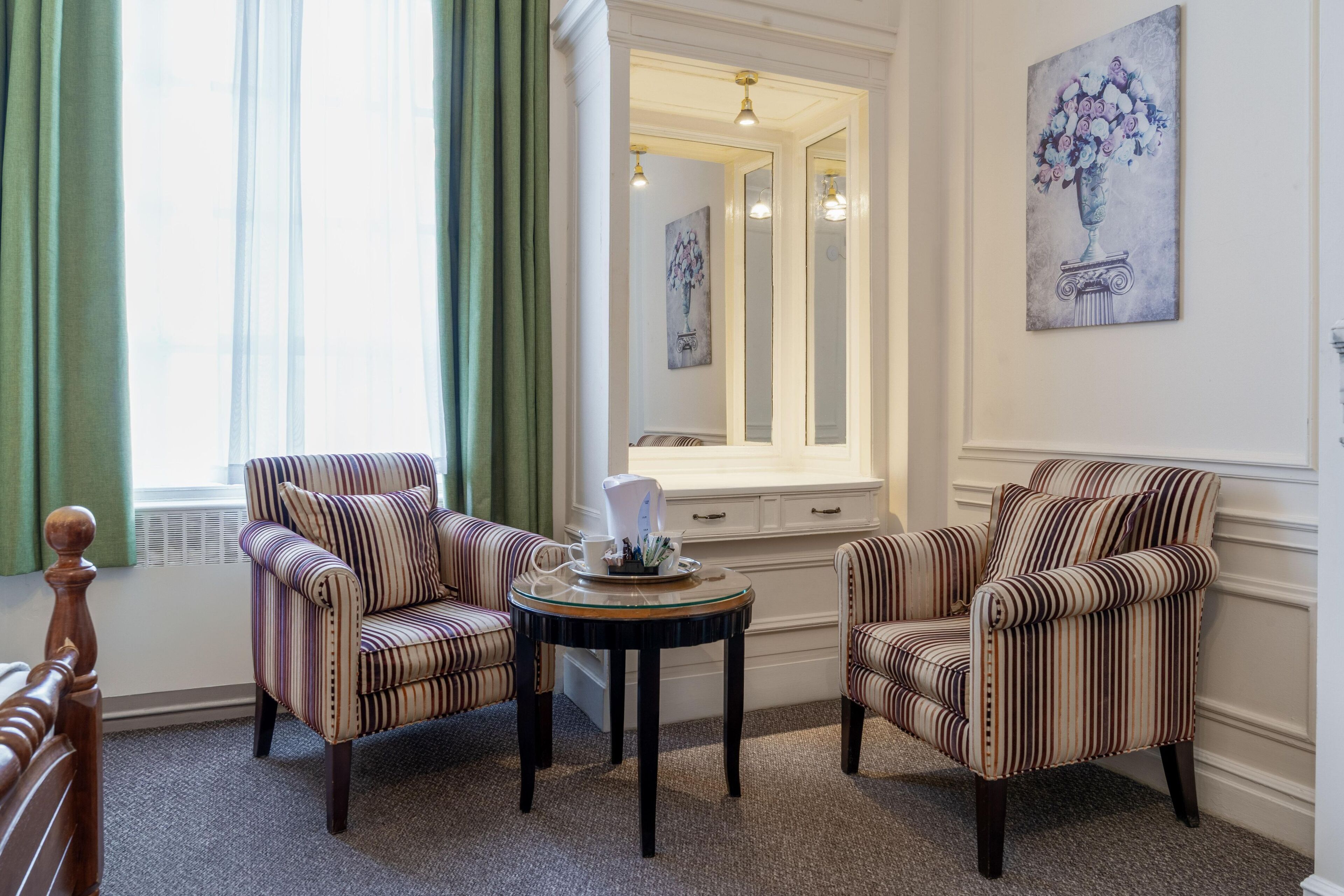 A room with two striped armchairs, a small round table with a white pitcher, and a large mirror reflecting the room.