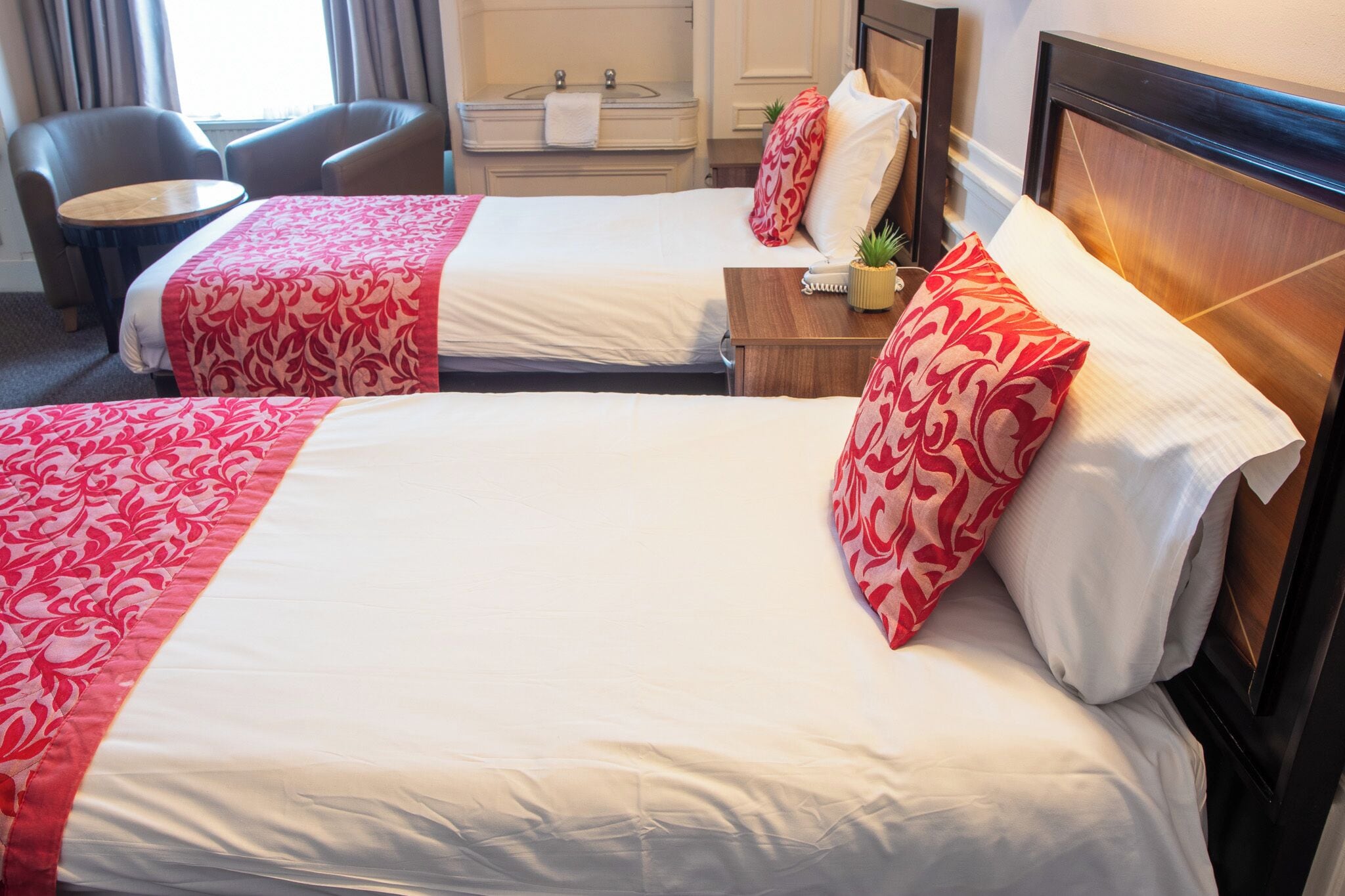 Una camera d'albergo con due letti, ciascuno con copriletto rosso e bianco a motivi geometrici, una testata imbottita in legno, un comodino con una lampada e una sedia.