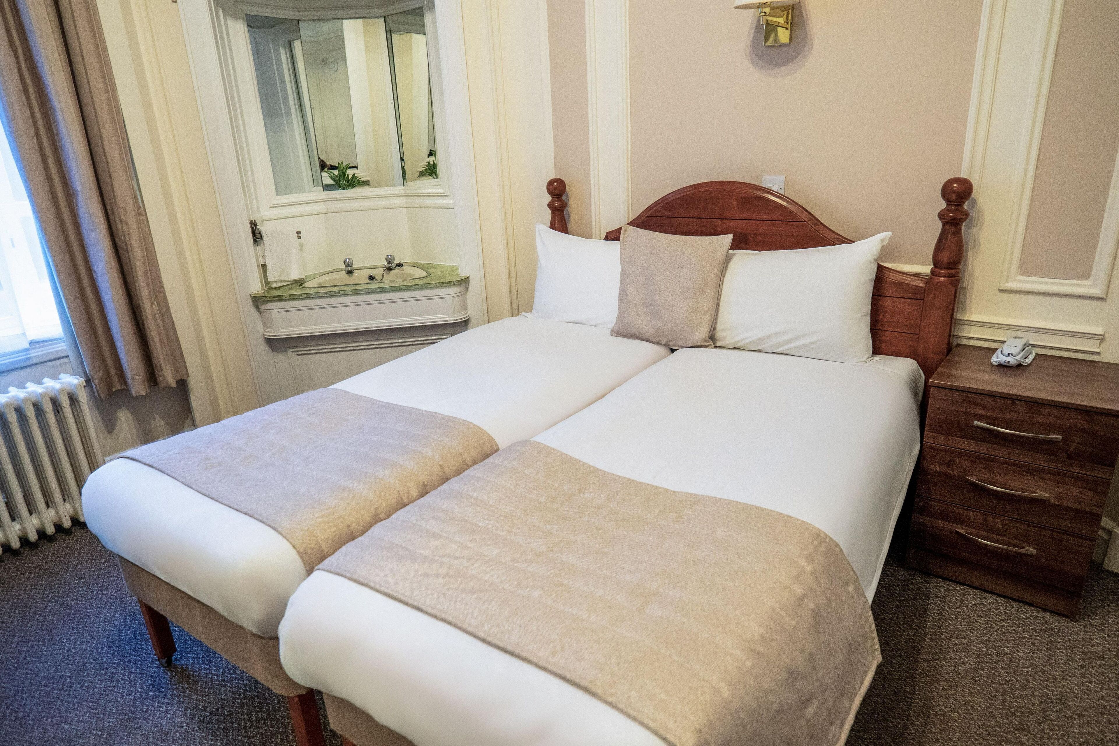 Una cama doble con dos almohadas, cabecera de madera, mesita de noche con lámpara y espejo sobre el aparador.