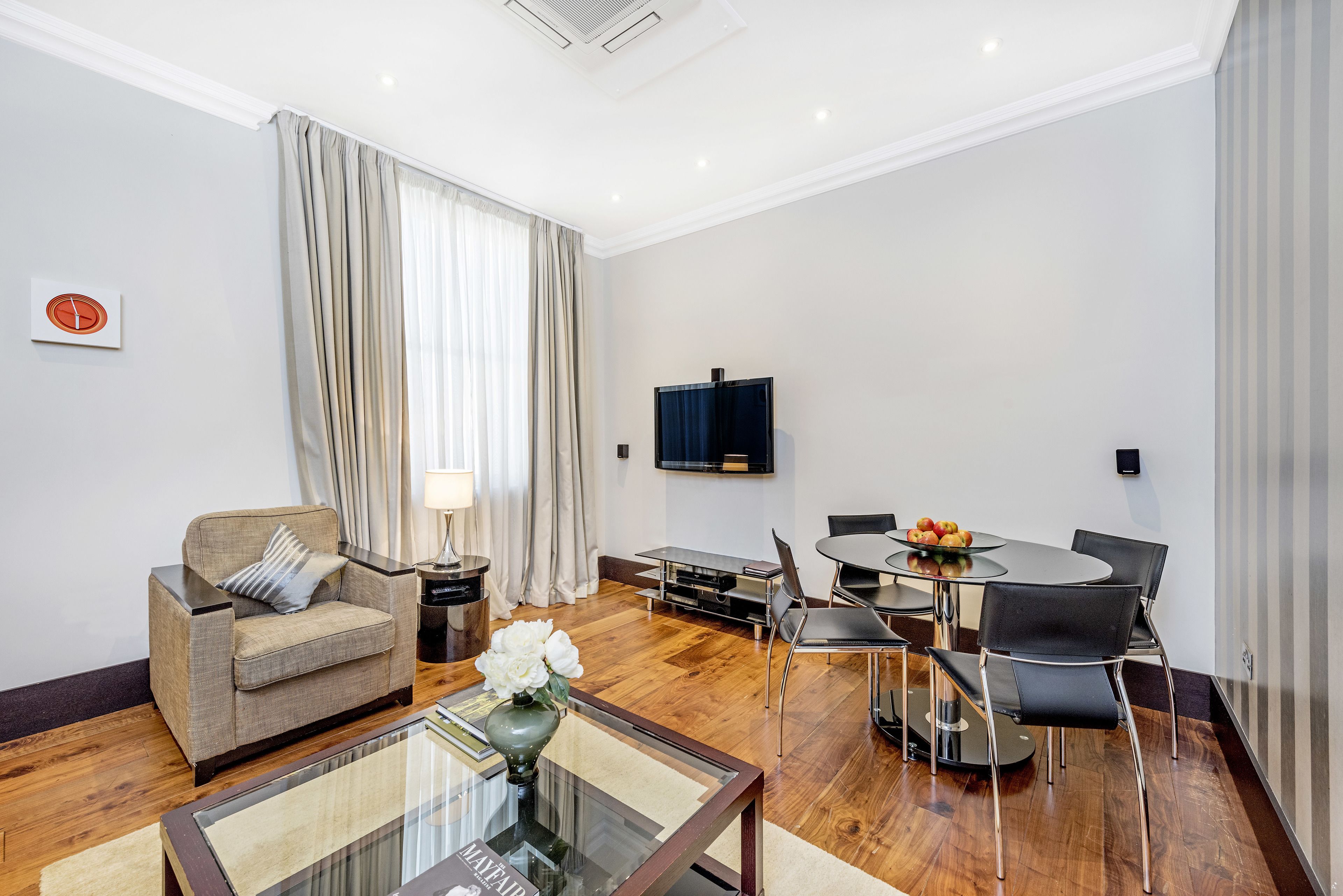 Una sala moderna con un sofá, una mesa de centro de vidrio, una mesa de comedor con sillas, un televisor de pantalla plana montado en la pared y un amplio ventanal con cortinas.