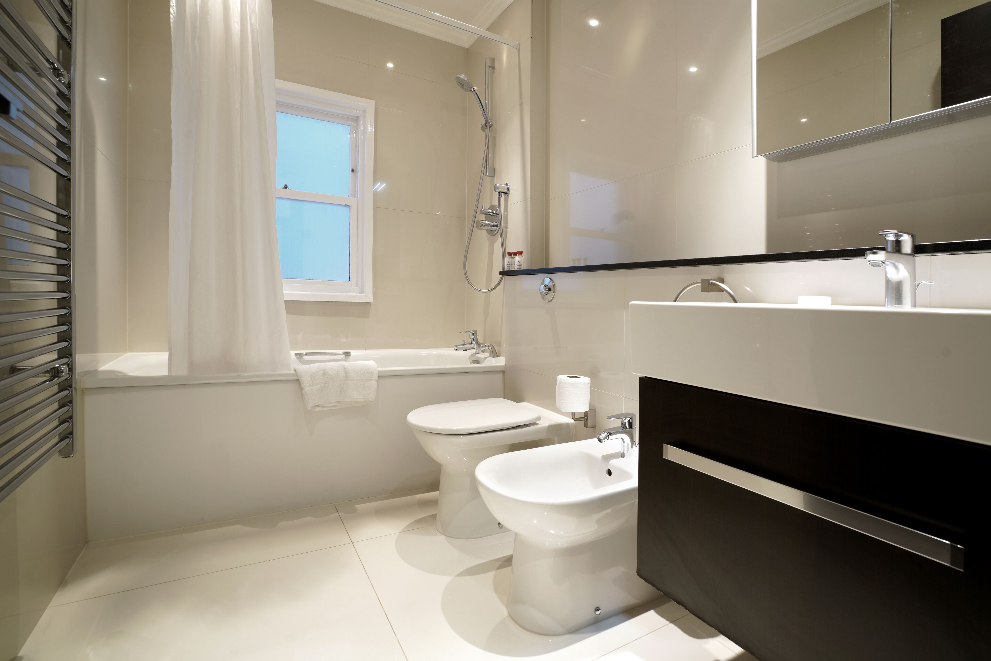 Et moderne badeværelse med badekar, to toiletter og en bidet samt et stort spejl.