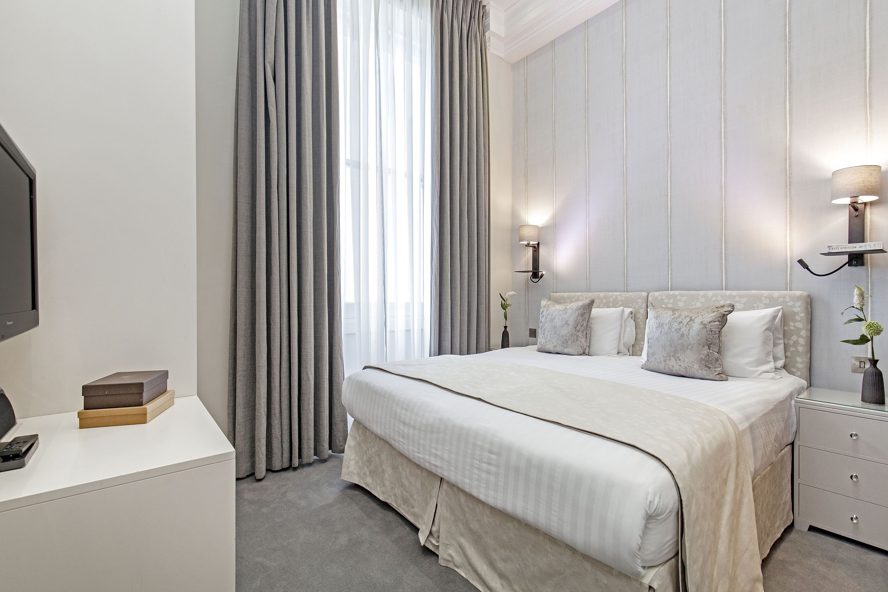 A modern bedroom with a large bed, bedside lamps, a TV, and a desk with a remote control.