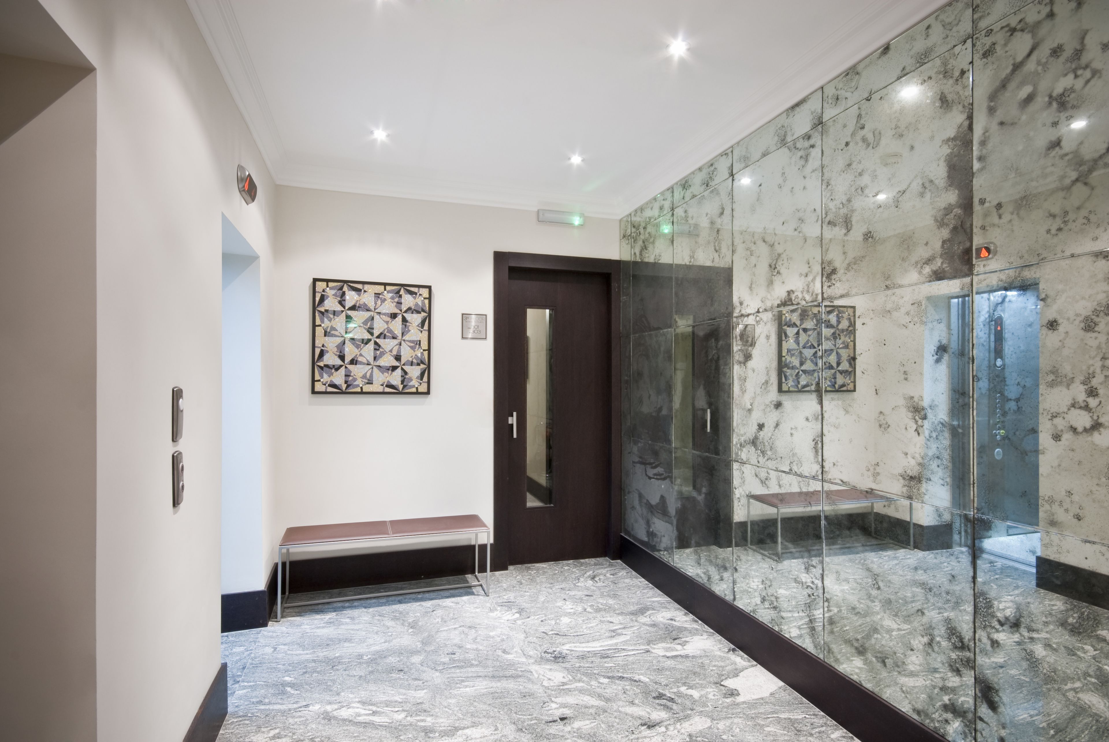 A modern hallway with a marble floor, a bench, a door, and a glass elevator.
