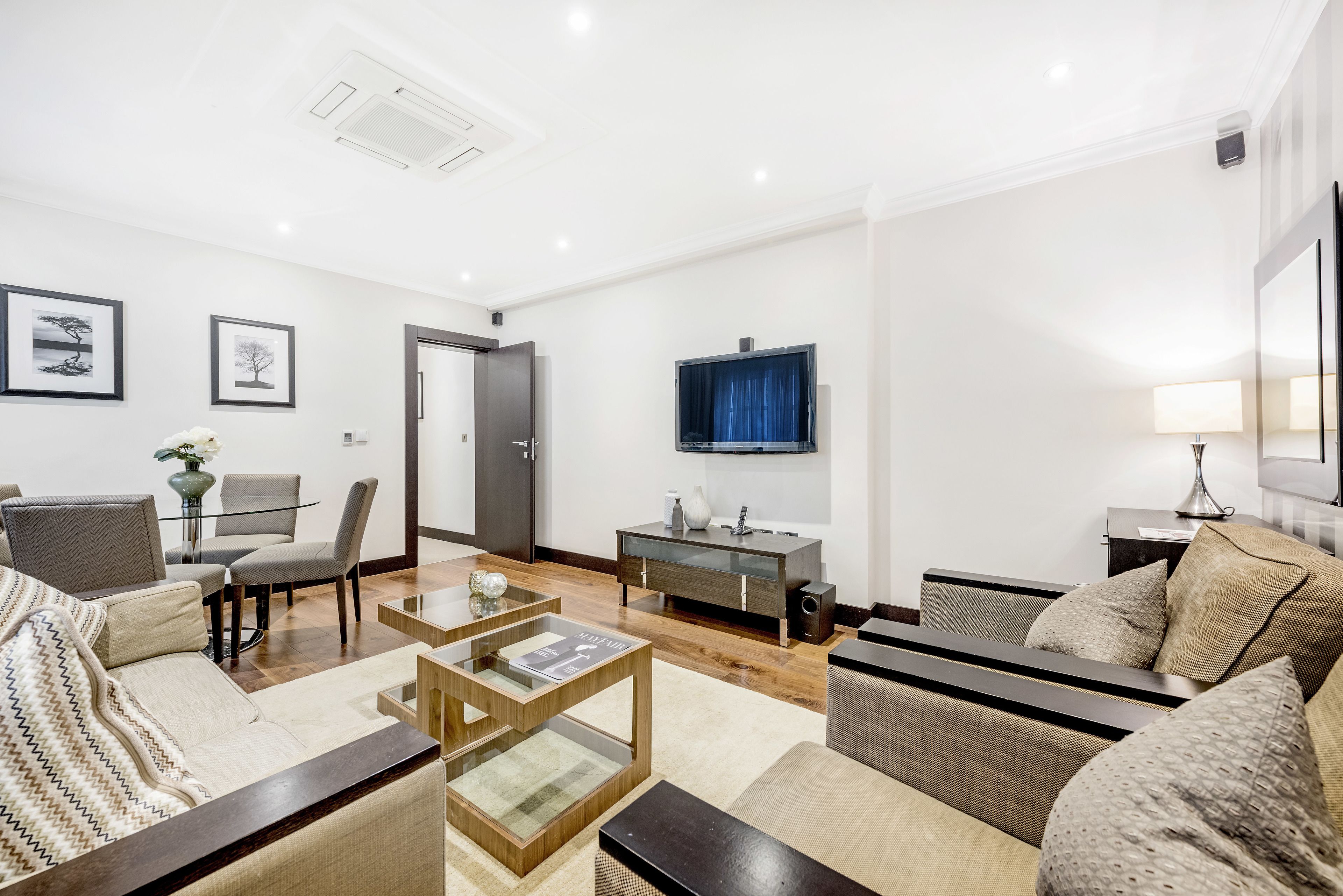 A modern living room with a sofa, armchairs, a coffee table, and a TV. There is a dining table with chairs and a small side table with a lamp.
