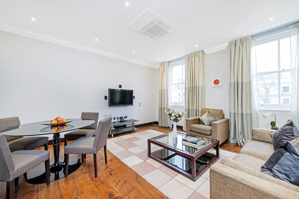 A modern living room with a glass coffee table, a round dining table with chairs, a sofa, and a television.