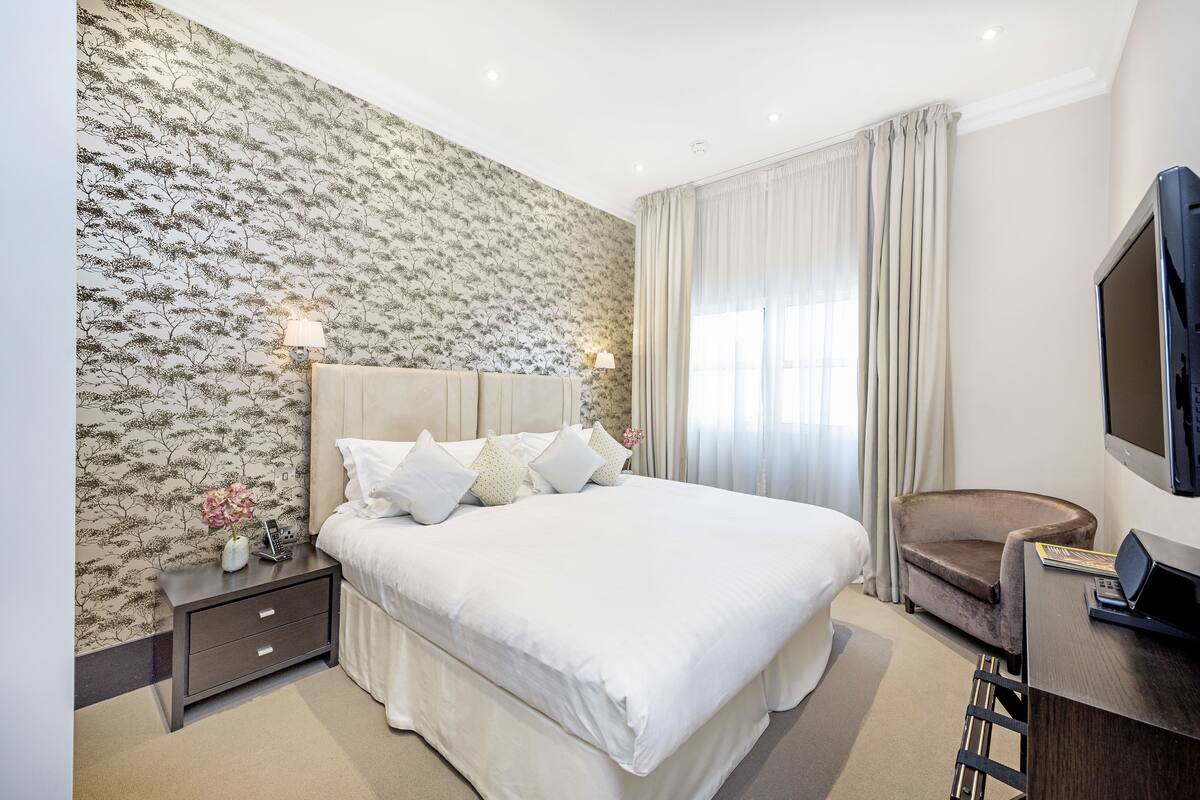 A neatly arranged bedroom with a large bed, a bedside table with a lamp, a chair, and a TV.