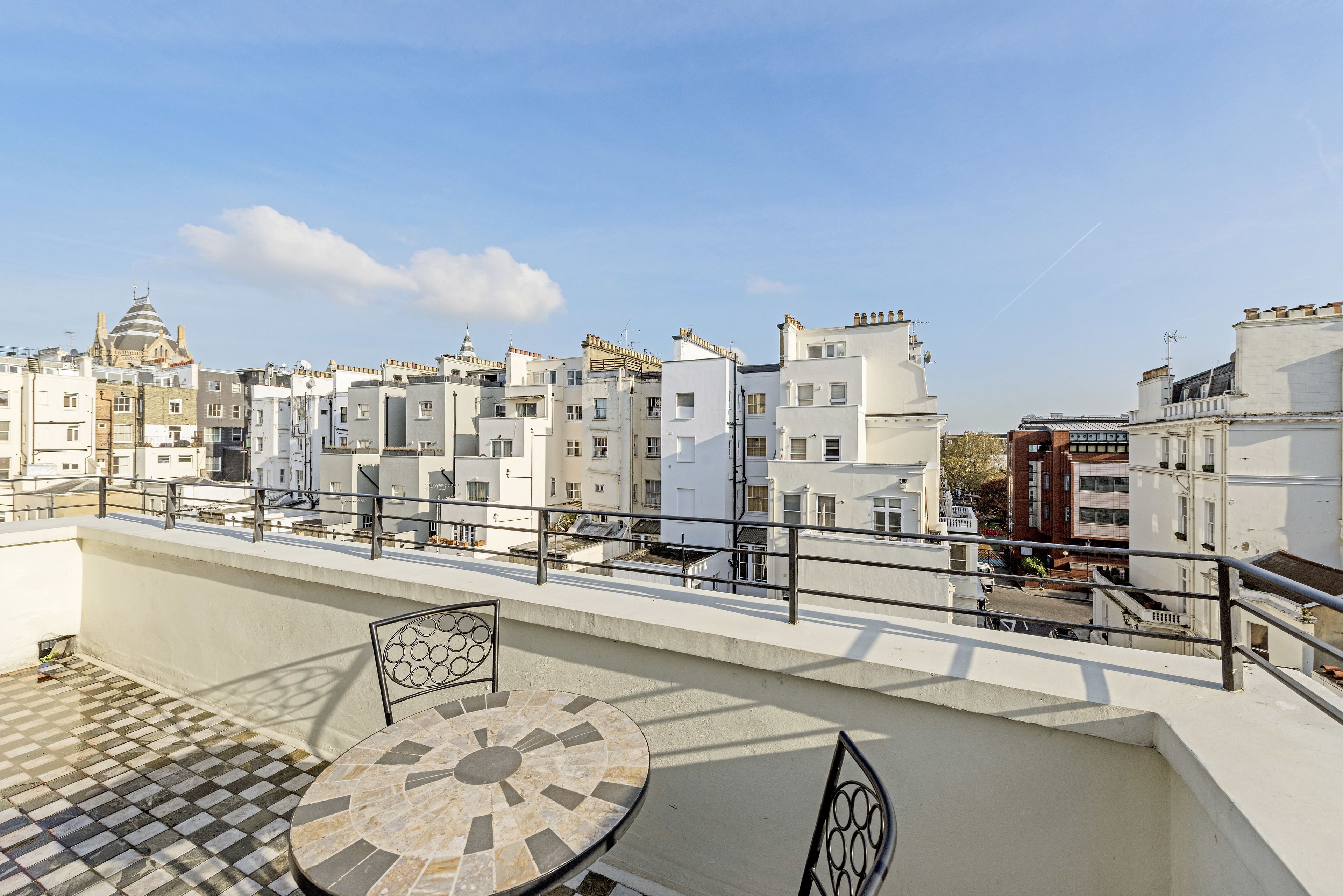 A rooftop terrace with a round table and chairs, overlooking a row of white buildings with varied architectural styles.