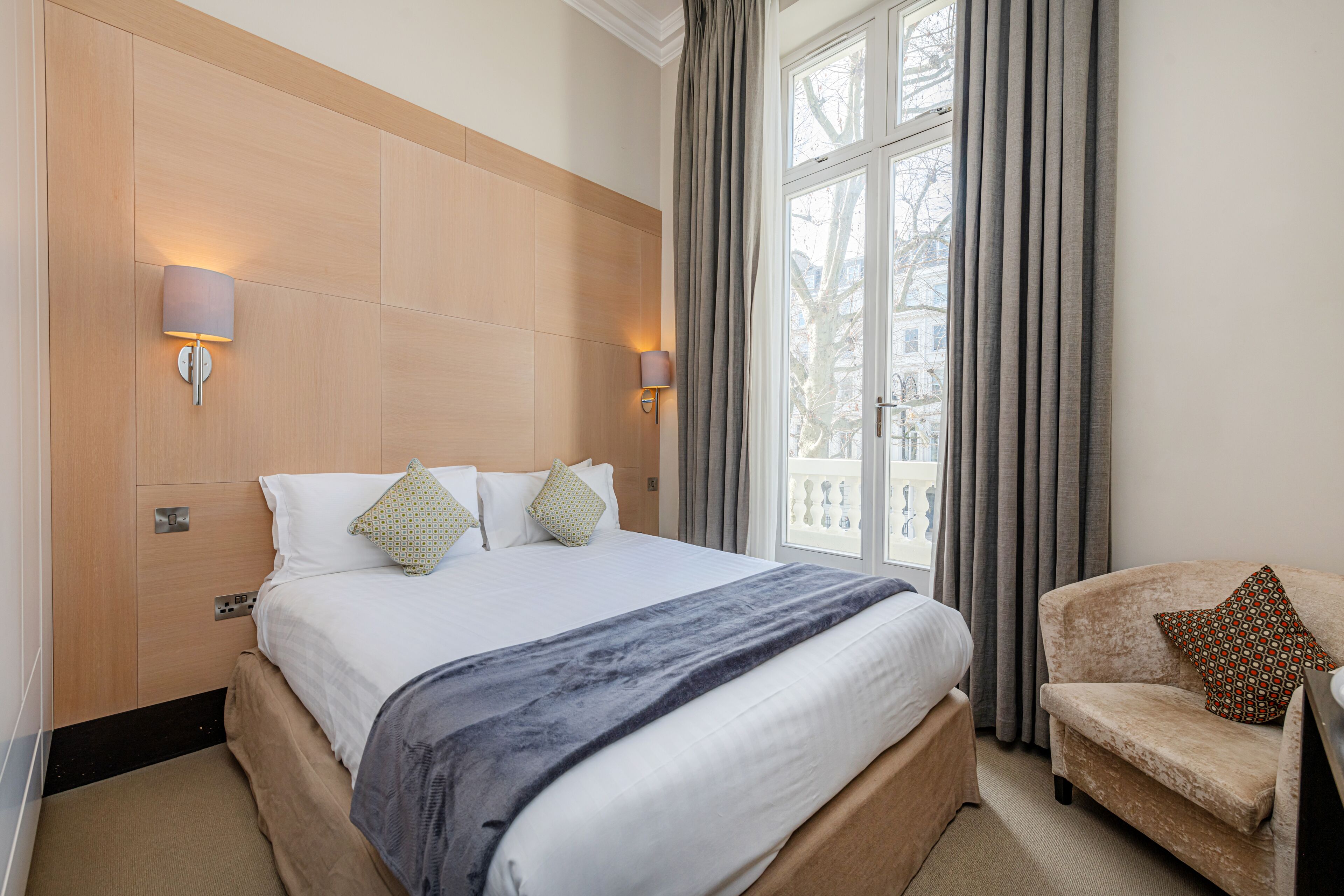 A hotel room with a large bed, two wall-mounted lamps, a window with curtains, and a chair.