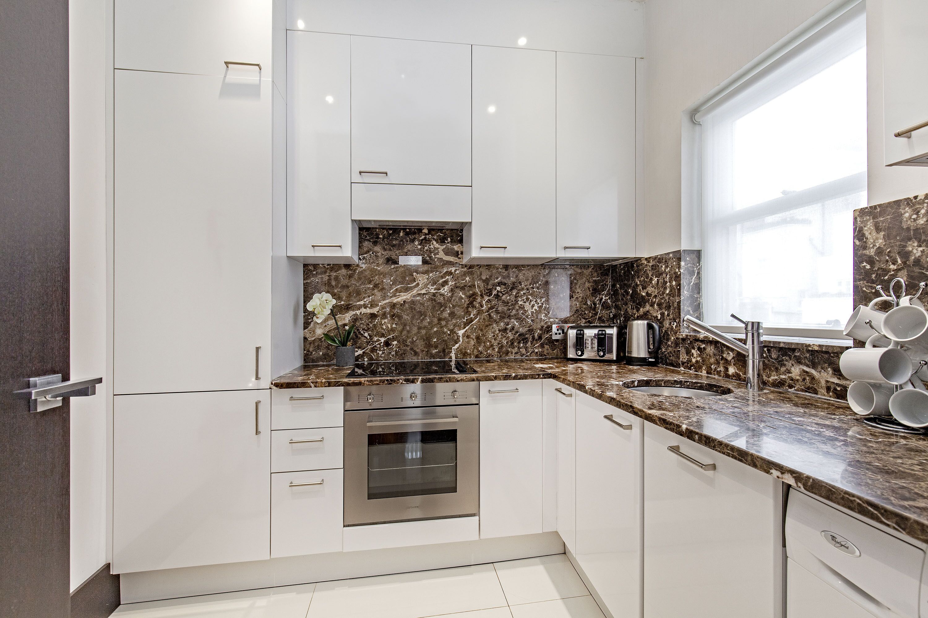 A modern kitchen with white cabinets, a stainless steel oven, a marble countertop, and a window providing natural light.