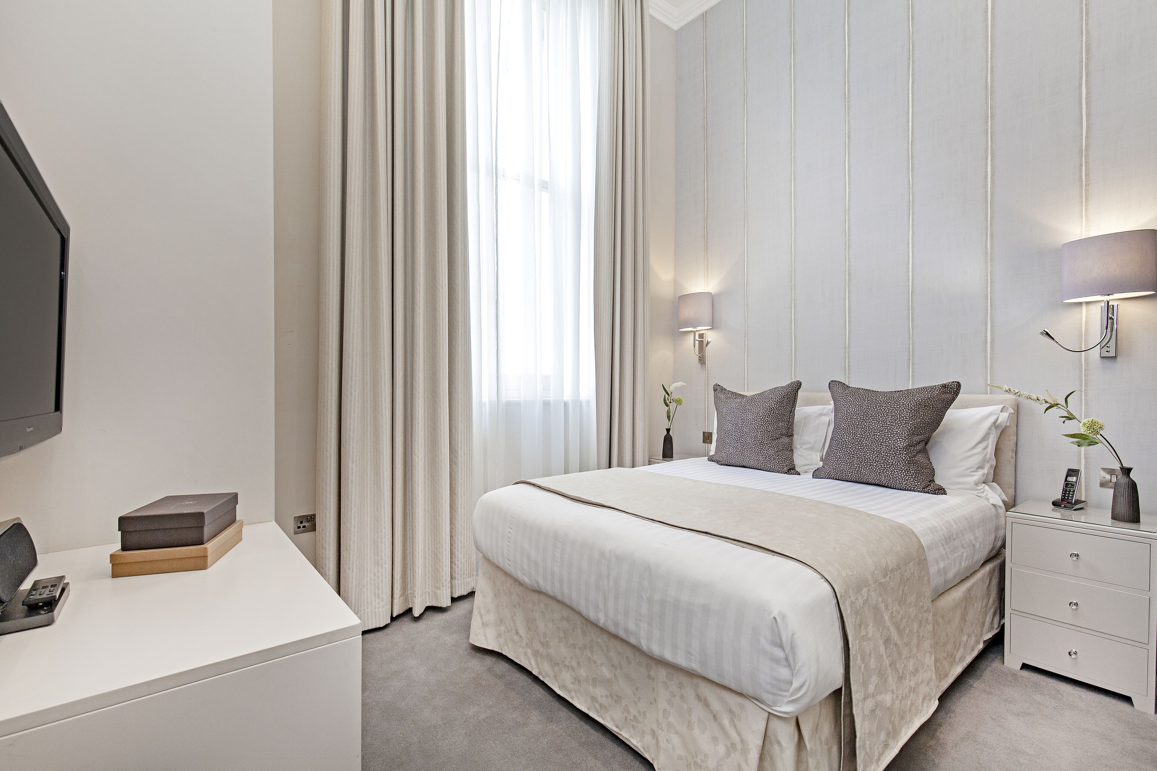 A modern hotel room with a large bed, bedside lamps, a desk with a phone, and a vase with flowers.