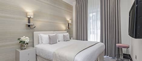 A modern hotel room with a large bed, two bedside tables, a flat-screen TV, and a view of the outdoors through sheer curtains.