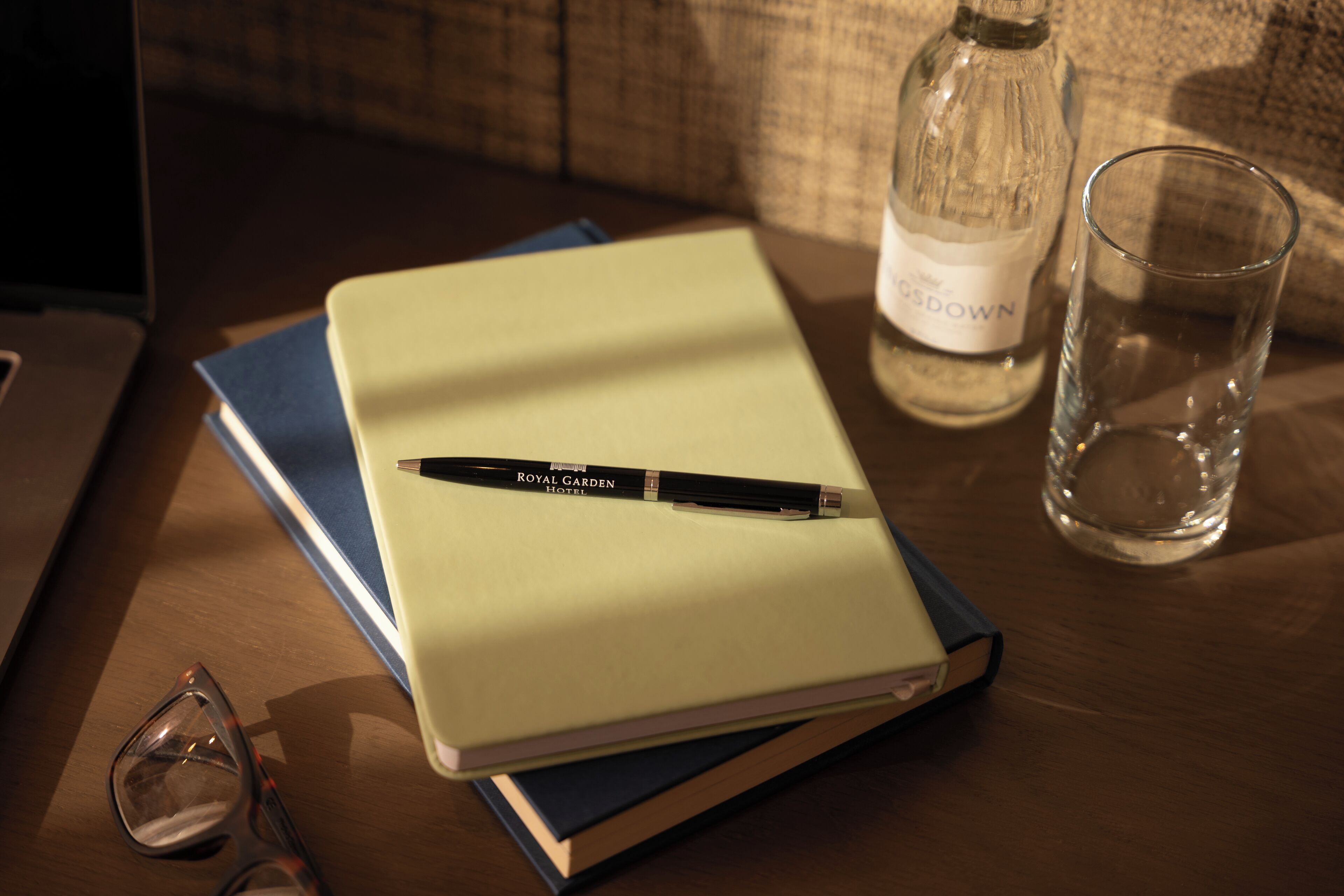 A notebook with a pen on top, a glass of water, and a bottle on a wooden table.
