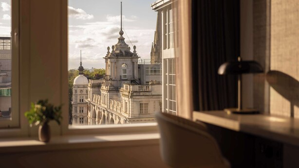 A room with a view of historic buildings, a window with a plant, and a lamp on the side table. - Royal Garden Hotel (London)