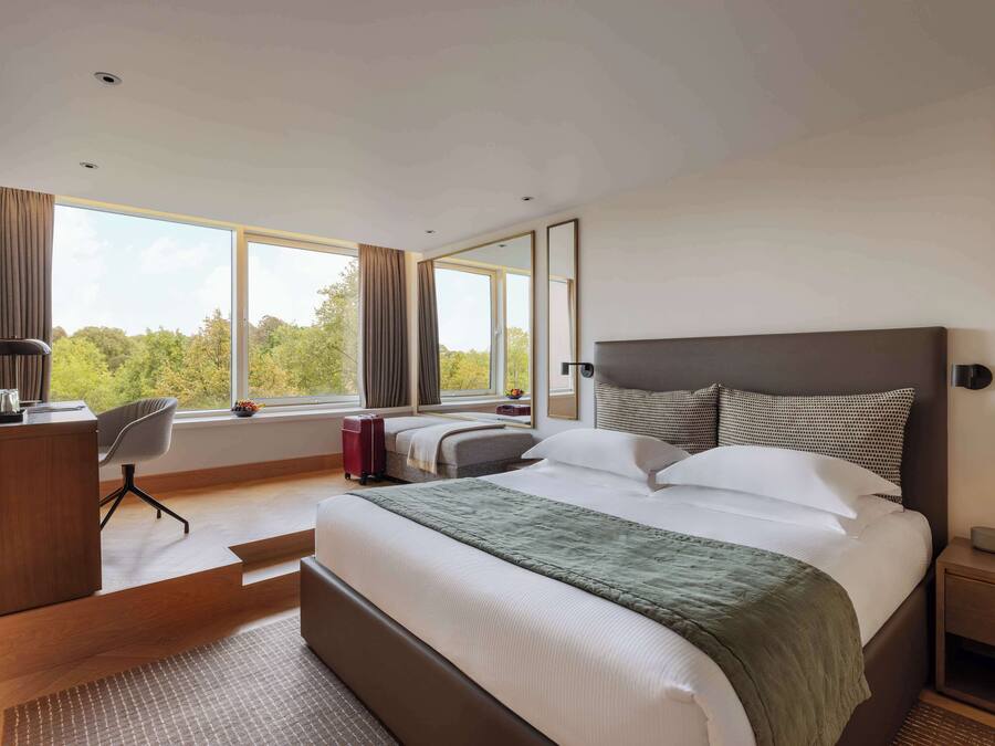 A modern hotel room with a large bed, a desk, a chair, and a view of trees through the window.