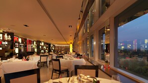 A restaurant with tables set for dining, a view of the cityscape through large windows, and shelves with decorative items. - Royal Garden Hotel (London)