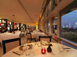 A restaurant with tables set for dining, a view of the cityscape through large windows, and shelves with decorative items. - Royal Garden Hotel (London)