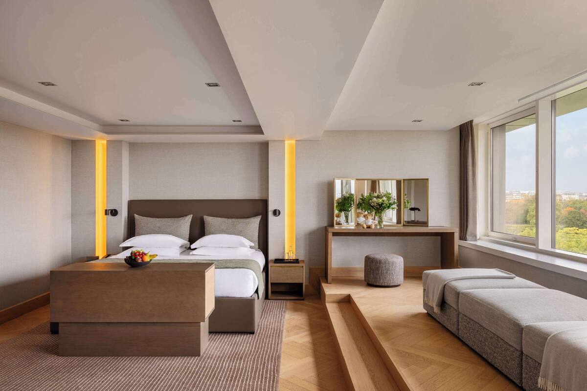 Une chambre moderne avec un grand lit, des tables de chevet, un bureau avec un miroir, un banc et une vue sur la ville.
