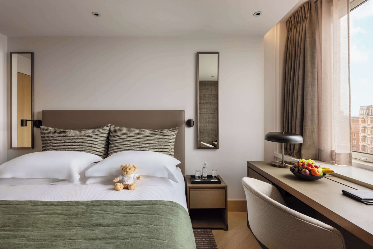A hotel room with a bed, a desk, a chair, a nightstand, a mirror, and a window with curtains.