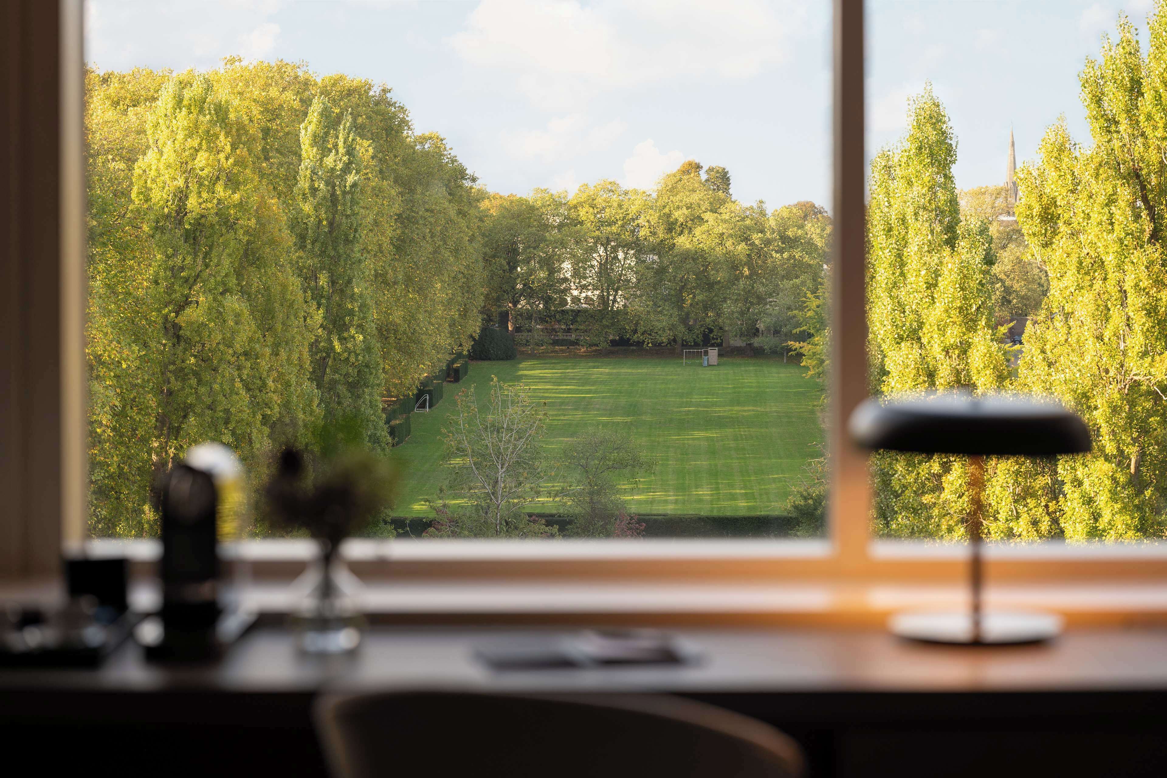 A desk with a lamp, a window, and a view of a green field and trees.