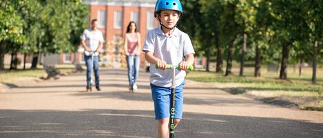 A child riding a scooter on a paved path with trees and a building in the background.