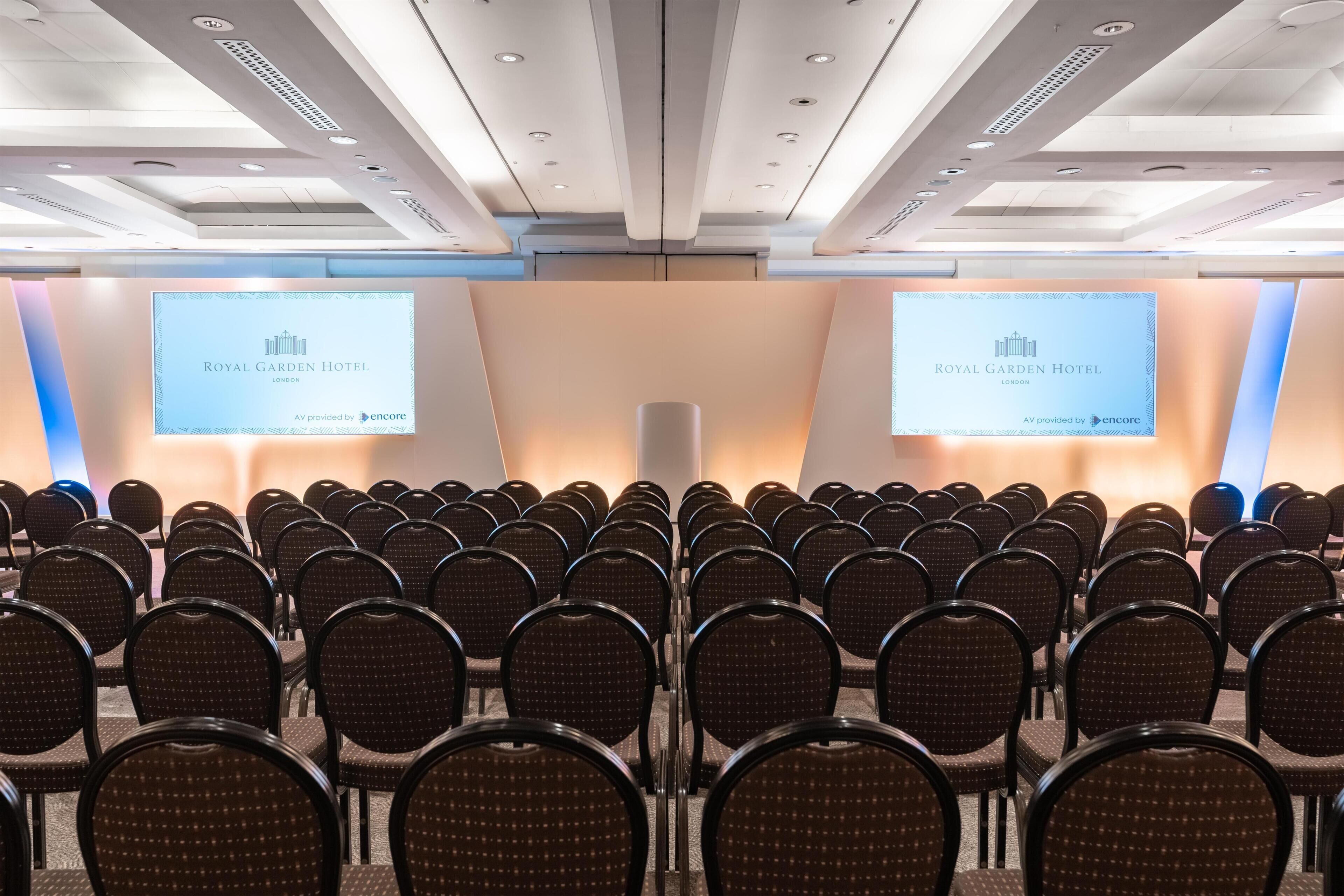 A conference room with rows of chairs, a podium, and two screens displaying the Royal Garden Hotel logo.
