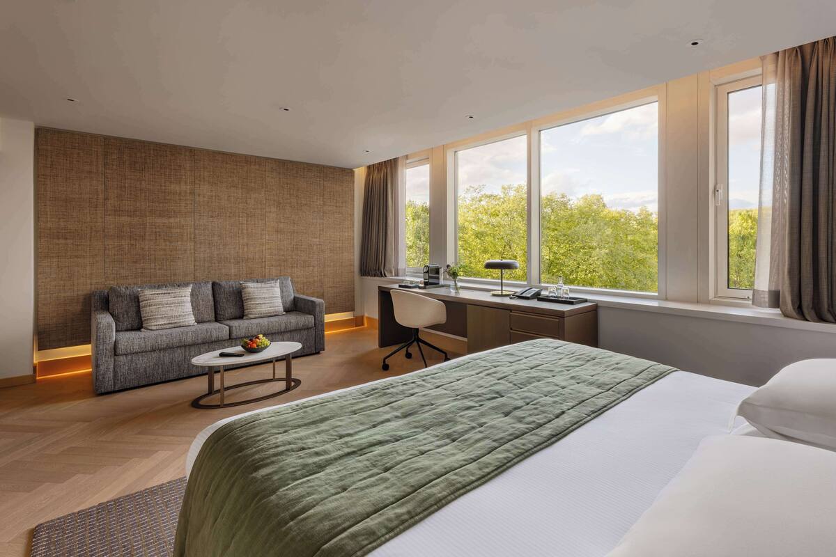 Une chambre d’hôtel moderne dotée d’un grand lit, d’un canapé, d’un bureau et offrant une vue sur les arbres grâce à de grandes fenêtres.