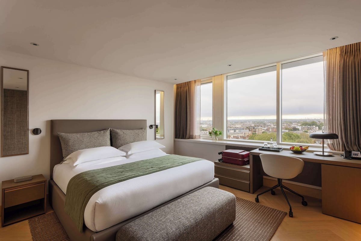 Une chambre d’hôtel avec un grand lit, un bureau, une chaise, une fenêtre donnant sur la ville et une petite table avec une plante.