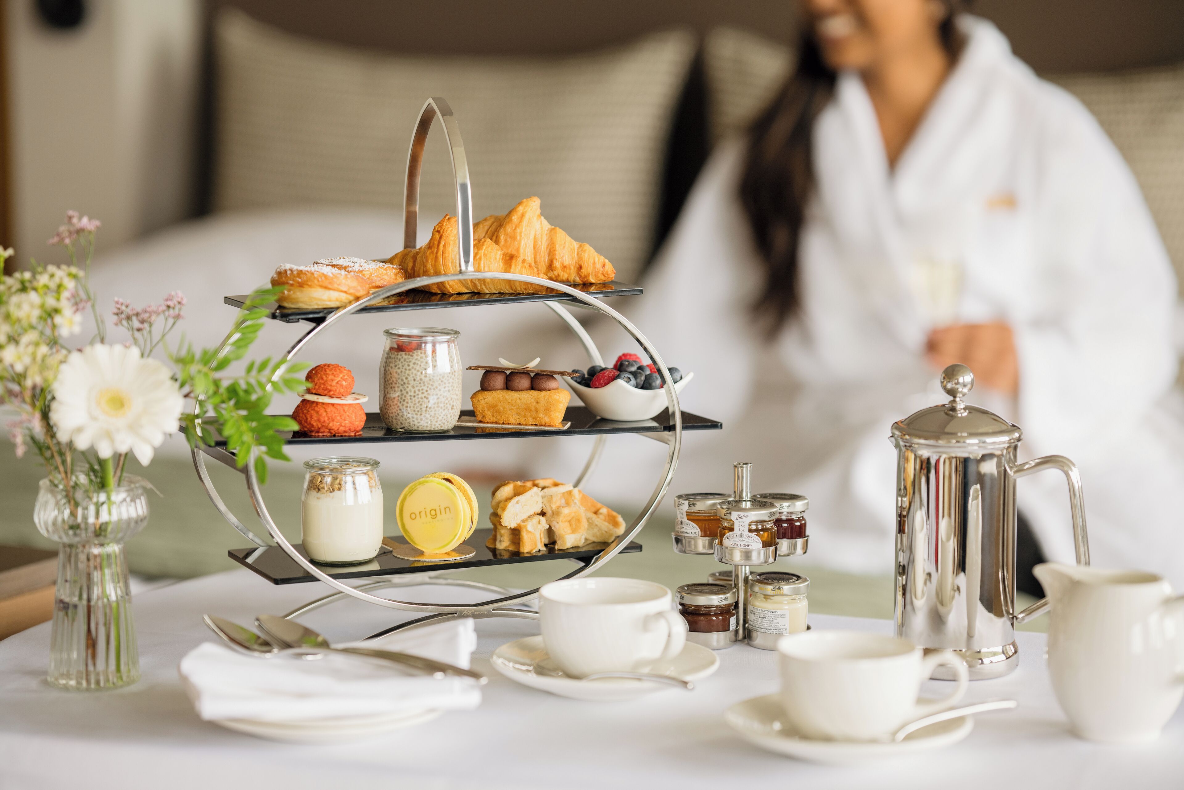 A breakfast setup with a three-tiered tray of pastries, a coffee pot, and various condiments on a table.