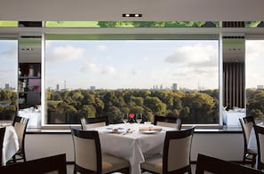 A dining area with a table set for two, large windows offering a cityscape view, and a bookshelf to the left.