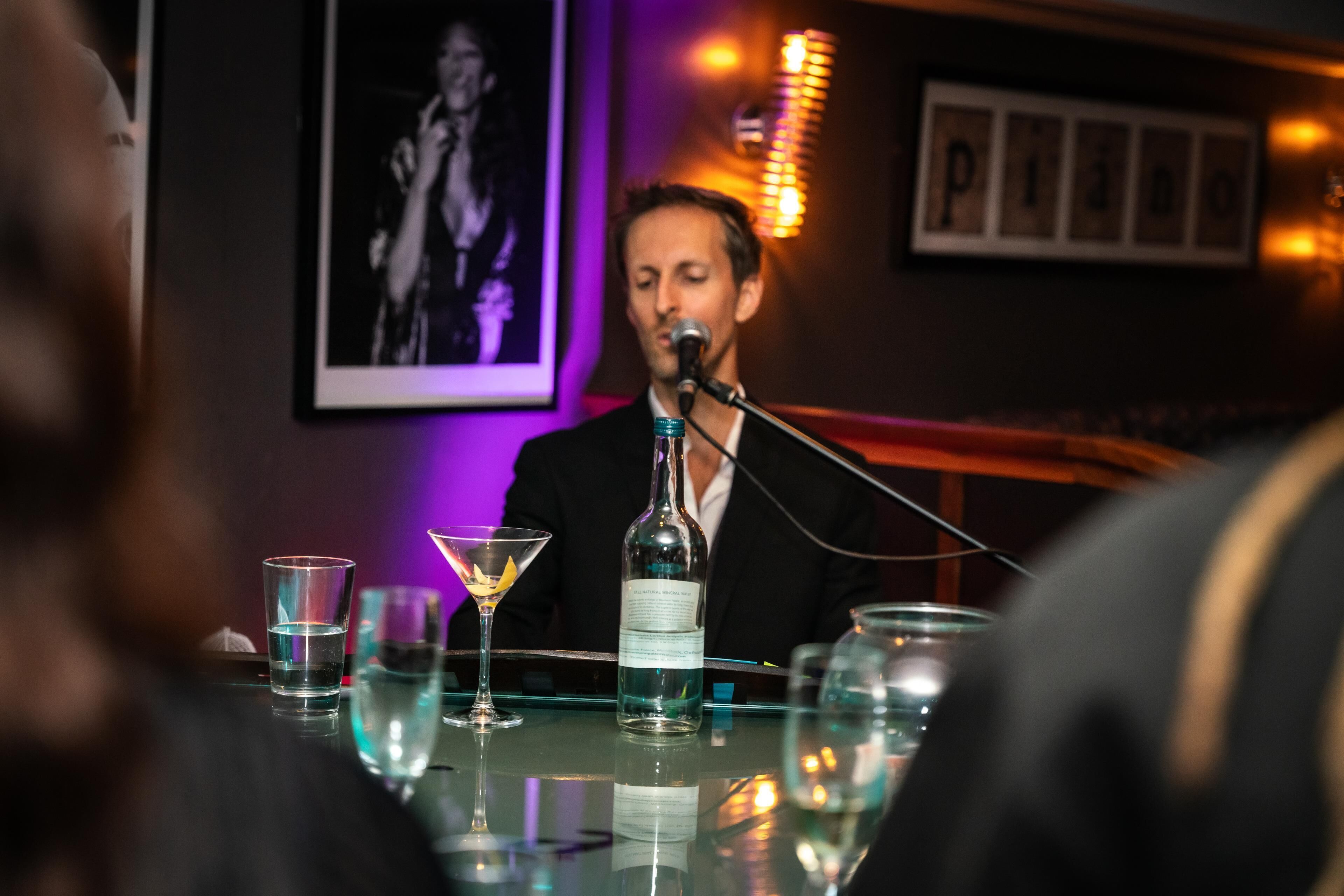 A person seated at a table with a microphone, a bottle, and glasses.