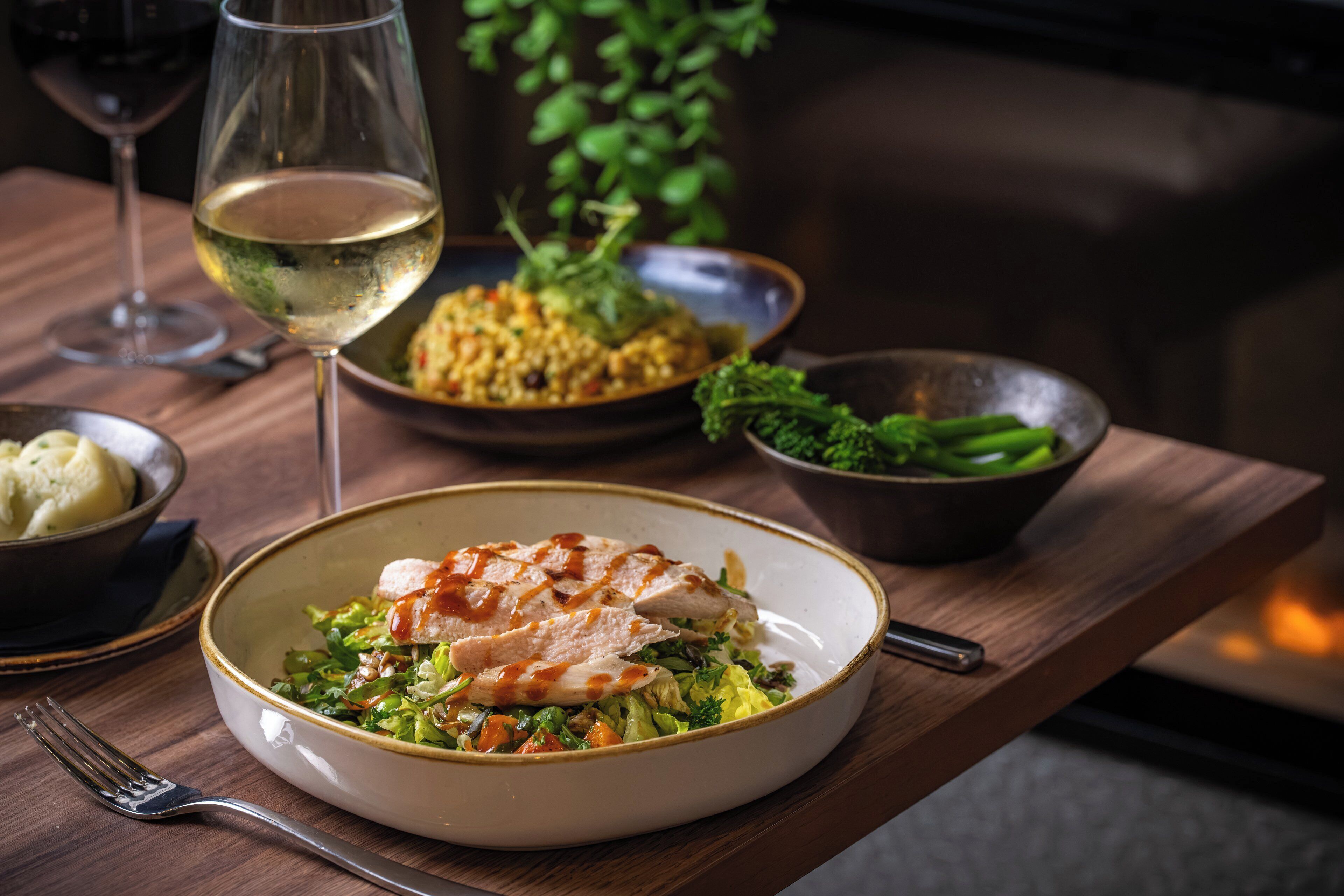 A dining table with a plate of food, a glass of wine, and a bowl of vegetables.