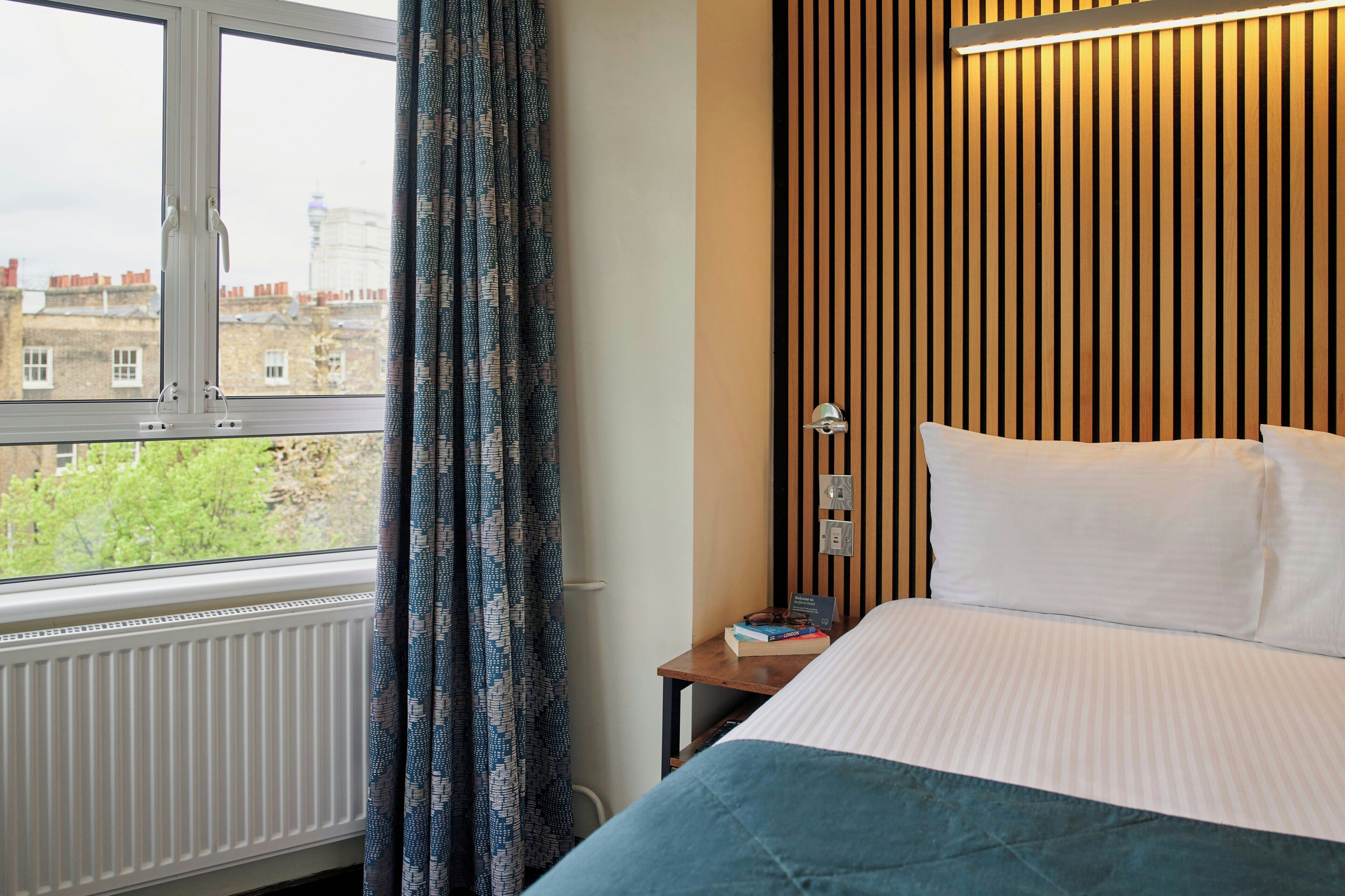 A hotel room with a large window, a bed with white bedding, a bedside table with a book, and a wall with vertical wooden slats.