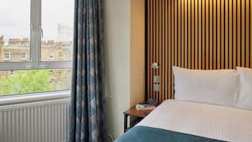 A hotel room with a large window, a bed with white bedding, a bedside table with a book, and a wall with vertical wooden slats.