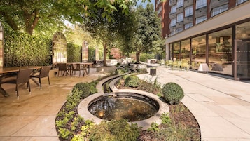 Ein Außenbereich mit einer Patio, einem runden Wasserspiel, Sitzgelegenheiten und einem Glasgebäude im Hintergrund.