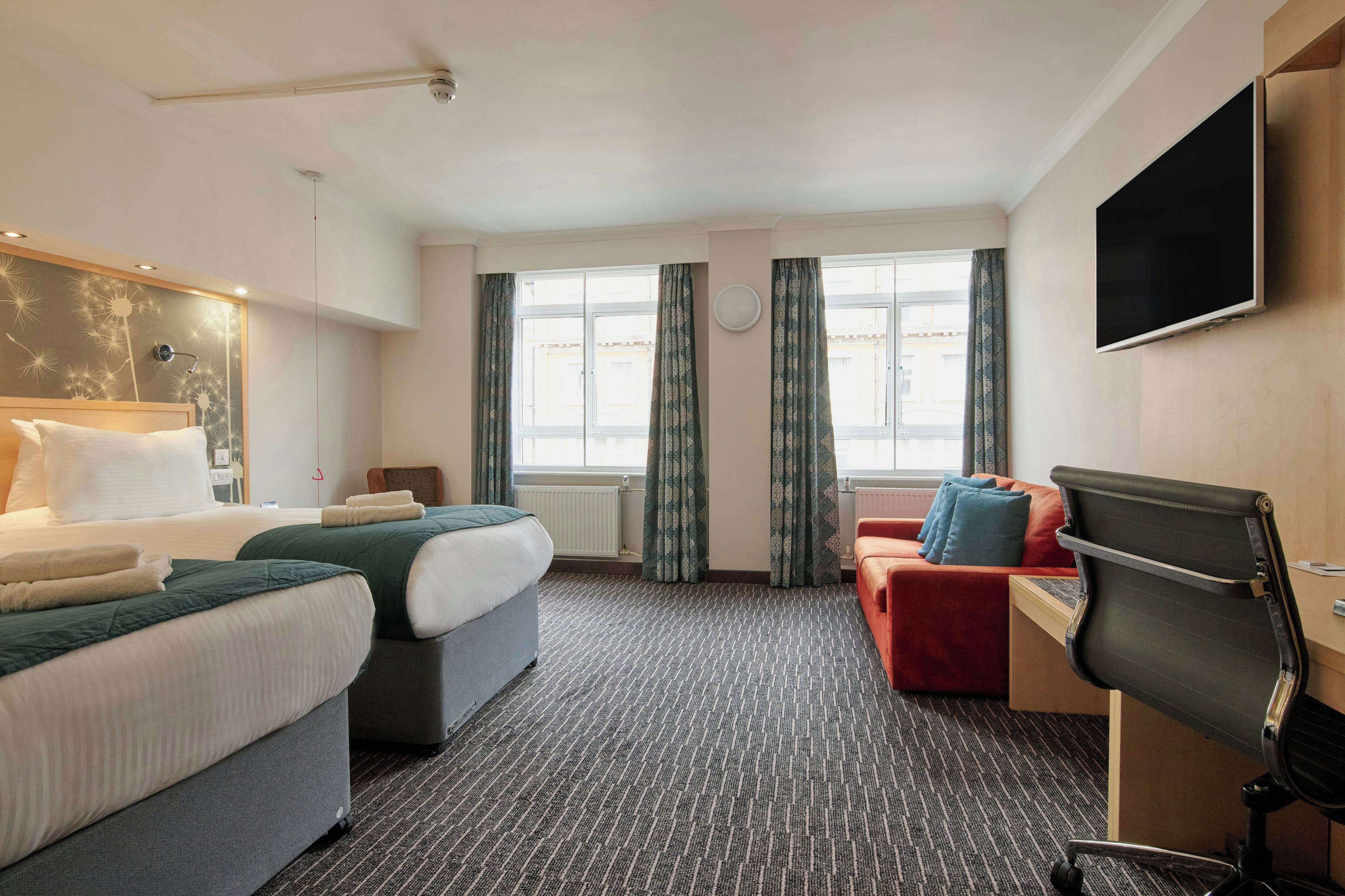 Ein Hotelzimmer mit zwei Betten, einem Schreibtisch, einem Stuhl, einem Fernseher und einem Fenster mit Vorhängen.