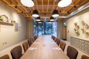 A conference room with a long table, brown chairs, pendant lighting, and a view through large windows.