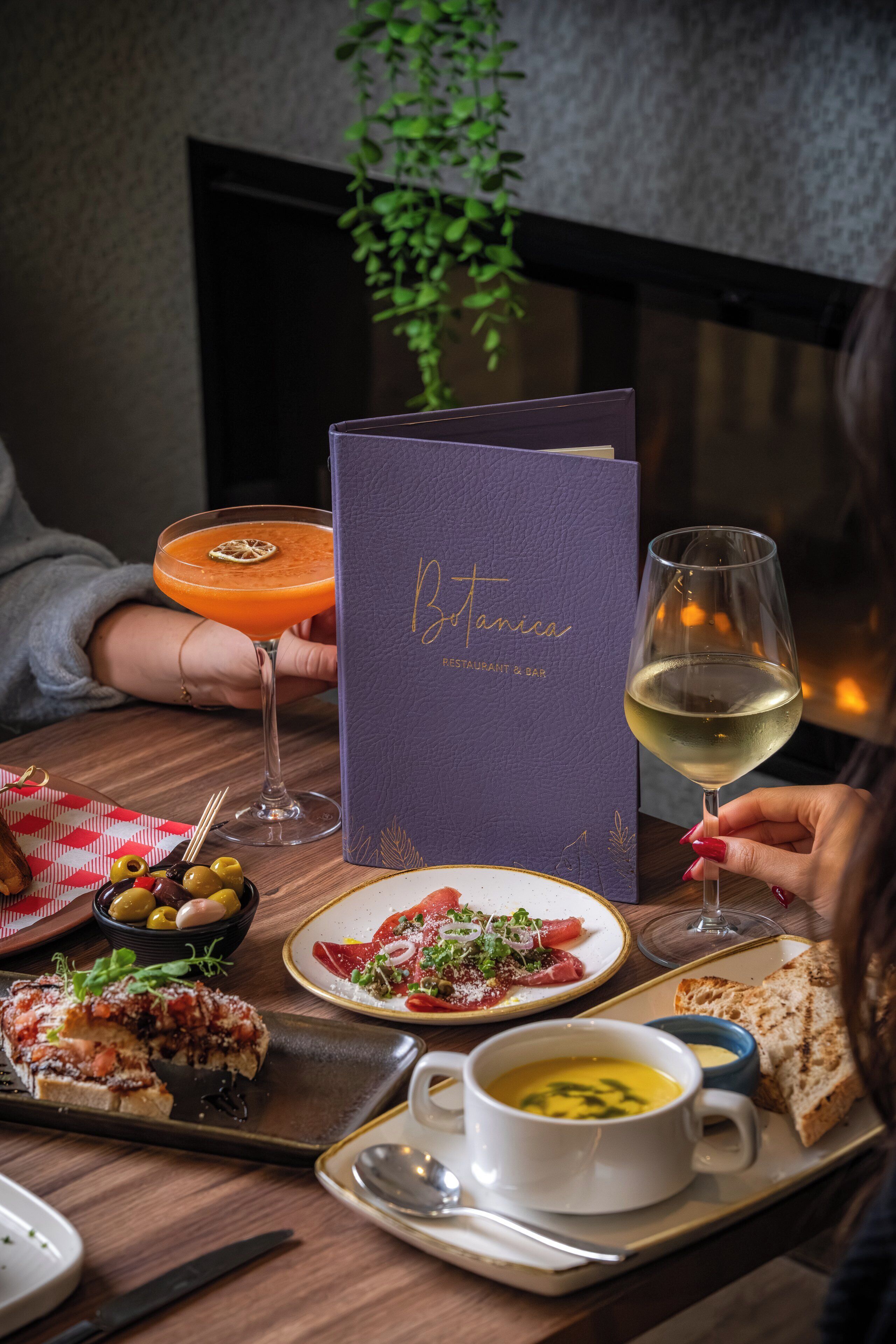 A dining table with a menu, a glass of white wine, a bowl of olives, a plate of food, and a soup bowl.