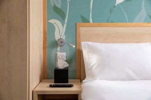 A neatly made bed with white sheets, a wooden headboard, a bedside table with a remote control, a tissue box, and a wall socket.