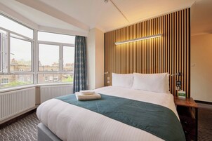 A hotel room with a large bed, a window with curtains, a wall-mounted light, and a radiator.