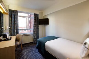 Ein Hotelzimmer mit Bett, Schreibtisch mit Stuhl, flachbildfernseher an der Wand und einem Fenster mit Vorhängen.